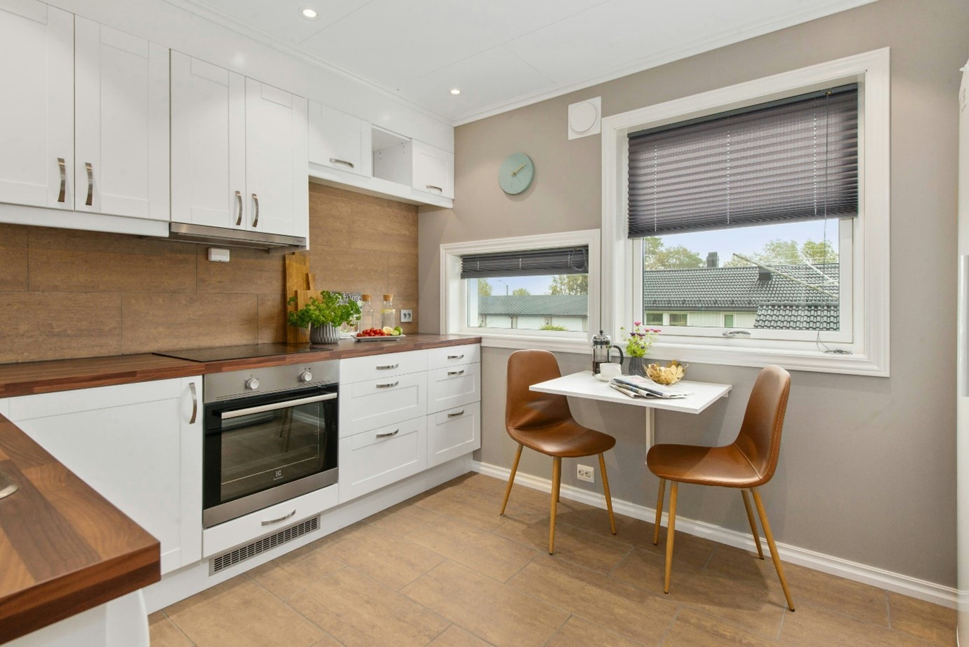 Cuisine blanche et bois avec éléments en hauteur, petite table et 2 chaises à côté d’une fenêtre