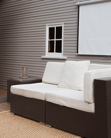 Sur une terrasse en bois, un salon de jardin en résine marron équipé de coussins blancs