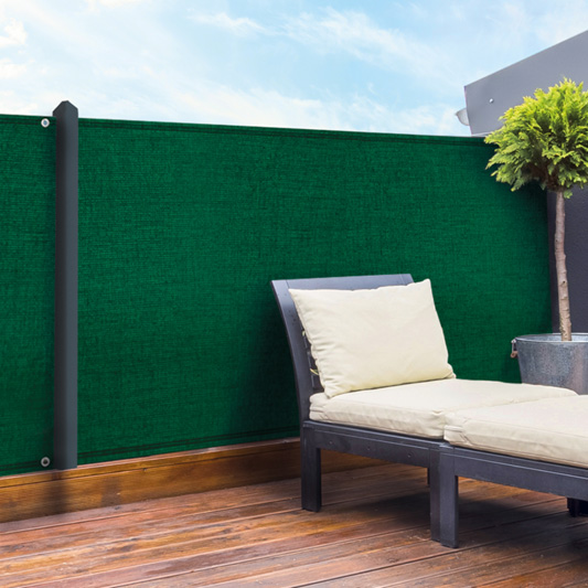 Sur une terrasse en bois, une clôture est installée avec un brise-vue vert et noir afin de protéger du regard extérieur le transat aux coussins blanc