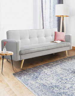 Dans un grand salon avec parquet, un long canapé gris clair semble moelleux et douillet. Il y a un coussin et un livre sur la banquette