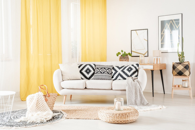 Salon dans les tons blancs, avec un canapé blanc et un pouf en osier. En arrière fond de longs rideaux jaunes