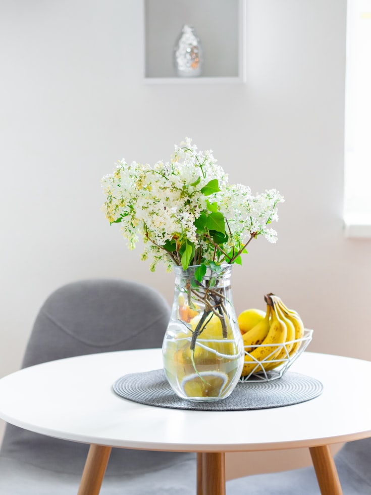 Table ronde blanche avec pieds en bois, chaises grises et vase de fleurs blanches au centre